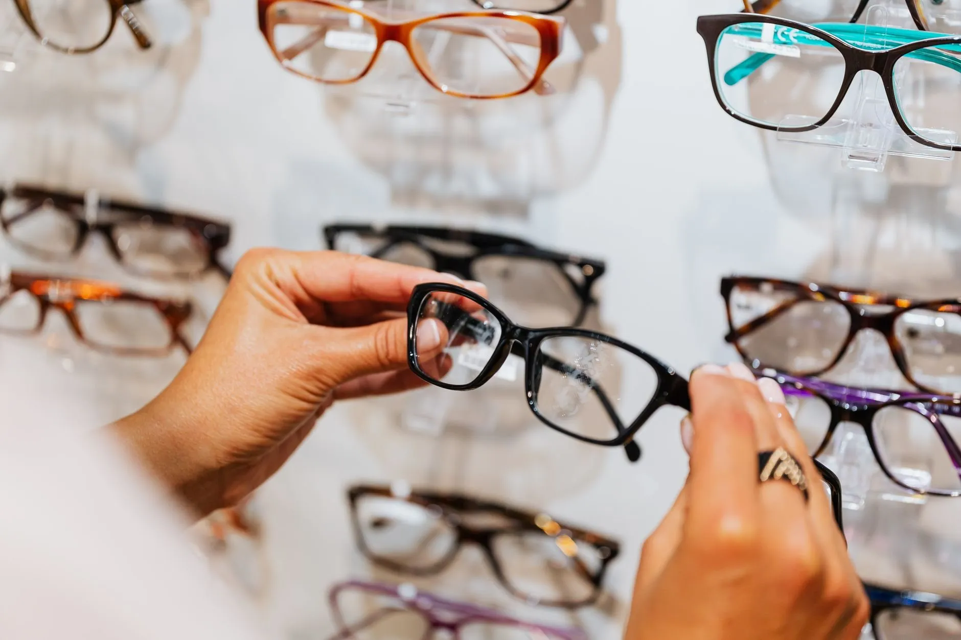 A hand holding a pair of black glasses up in front of a wall of frames