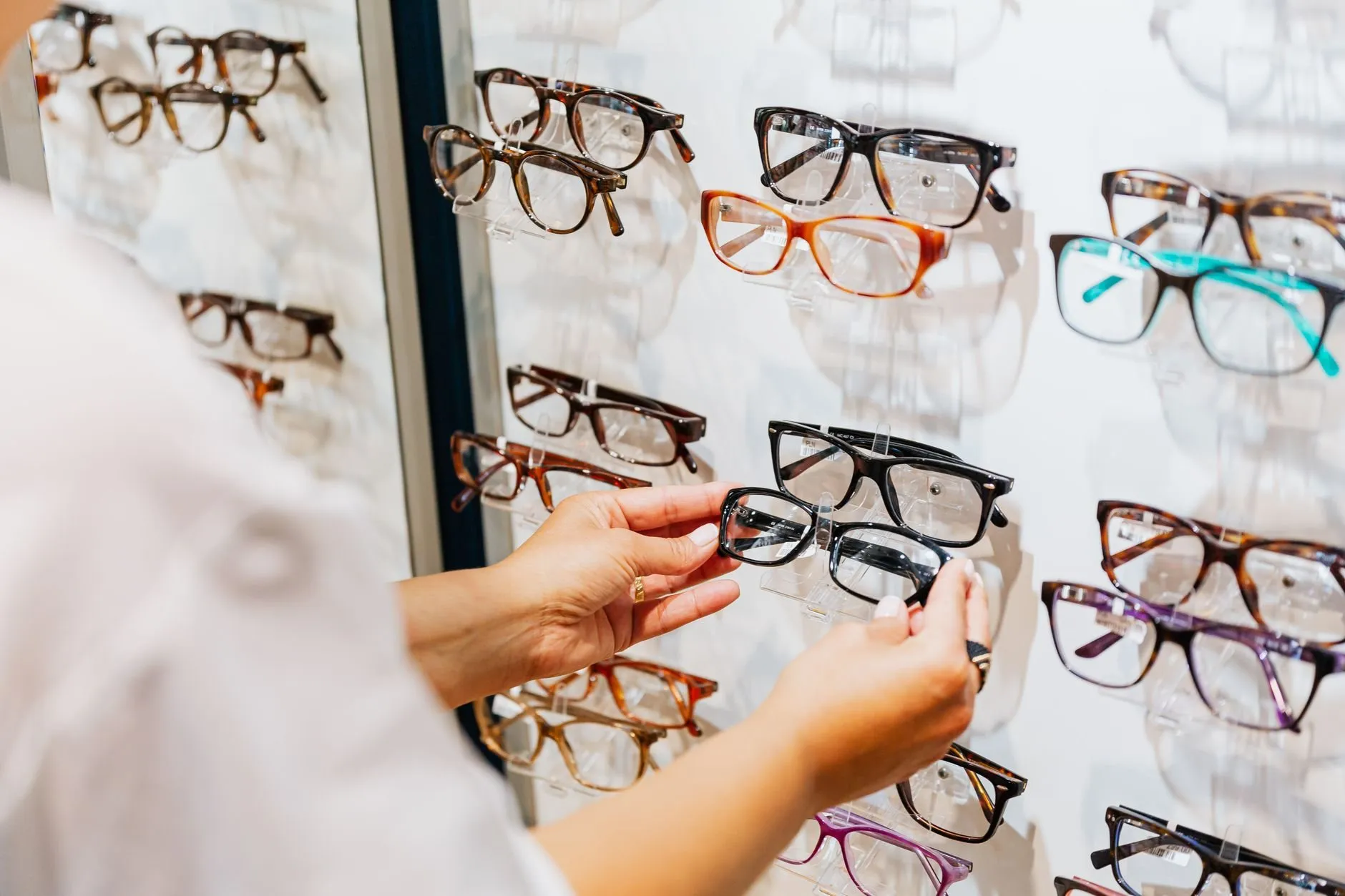 Hands taking a pair of black frames down from a bright wall of glasses